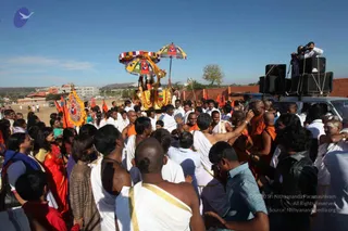 Outdoor procession on an open dry field under a clear blue sky. A crowd of men in white dhotis with gold borders and orange-robed monks surrounds a decorated palanquin carried on poles, topped with multicoloured striped canopies and garlanded with orange and yellow flower strands. Behind it, orange and red ceremonial flags flutter beside a large yellow-framed banner. A speaker stack and a white van stand at the right, with a cameraman on top filming. Women in saris and salwar kameez watch at the left edge.