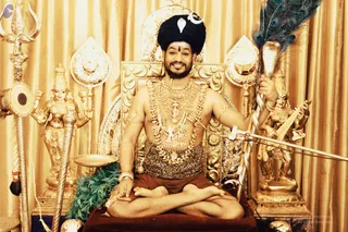 A bare-chested man sits cross-legged on a red cushion before an ornate gold throne against gold drapery. He wears a tall black turban with a white crescent and leaf ornament, a red bindi, and heavy layered gold necklaces with rudraksha and gold pendants. In his right hand he holds a peacock-feather fan, and in his left a stem of green leaves. Gold deity murtis flank him, including a trident on the left and a veena-playing figure on the right, with a gold kalasha-like tray nearby. a person: the bare-chested figure wears a turban, rudraksha and ceremonial gold necklaces, and sits on a throne, so the appearance-based label applies.