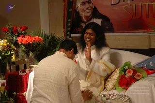 A photograph of an indoor event with a long-haired man in white ceremonial clothing seated on a white-draped bed or dais, smiling and raising his right palm in greeting while a man in a light checked shirt bends forward toward him. The seated man wears a white silk-like garment with gold border, a gold necklace, and a red bindi. Behind him hangs a red banner with a large portrait labelled "Nithyananda" and the word "SUTRAS" in gold letters. Bouquets of red and orange roses, yellow flowers, palm fronds and a bouquet of pink and red gerbera flowers fill the scene, with a red-draped table holding a candle and a plate of printed cards at the lower edge.