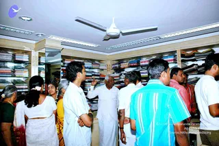 Photo inside a clothing showroom with floor-to-ceiling shelves stacked with folded garments in many colours, under a white ceiling fan and recessed lights. A group of about ten people crowds the aisle looking up toward an older man in white dhoti and white shirt who points upward with one hand. Nearby a woman in a white sari with a flower in her hair, a woman in a yellow sari, and men in white shirts and a turquoise striped shirt listen. The shelves are lined with saris and shirts, and a sign on the upper shelf reads Nithya.