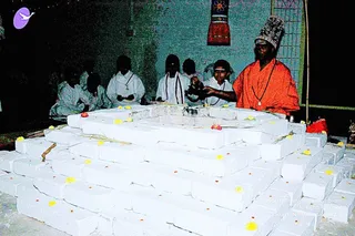 Night photograph of a ritual at an outdoor altar built from stacked white rectangular blocks, decorated with yellow and orange flower petals and a few small red flowers. A man in an orange robe and a tall patterned cap stands at the right, reaching toward the altar with a small object in his hands. Behind the altar, several young people in white clothing sit on the ground, some with white garments and a cord across the shoulder, one holding a small item. A red cloth hangs on the wall behind them, and a sheet of yellow garlands and incense sticks lies on the altar. The setting appears to be a Navaratri observance at Aadi Kailaasa in Bengaluru.