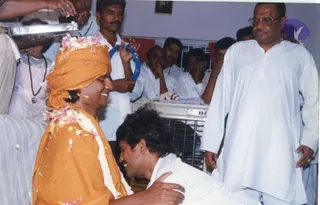 A photograph of an indoor gathering where a man in an orange-saffron robe and orange head wrap, decorated with pink flower petals and a white garland, smiles and places a hand on the shoulder of a man in white who bows low with his head down. Scattered flower petals lie around them. A man in white kurta and round glasses stands at right, and seated devotees in white clothes watch, some holding a blue tambourine. A framed picture hangs on the wall above and a metal cage-like grill is behind. Appearance-based candidate with saffron robe, orange head wrap and garlanded ceremonial dress: a person, receiving a bowing devotee in an energy darshan setting.