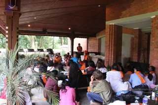 A covered open-air hall with brick pillars and a wooden ceiling, where a seated audience of devotees gathers in front of a low stage. Many attendees wear orange shawls or stoles and sit on the floor and on wooden chairs, some in pink, blue and striped clothing. At the front, a man in an orange robe stands near a figure in an orange turban and orange clothing seated at a white-draped table with flowers, which suggests a satsang or discourse. A potted palm sits in the foreground beside a camera on a tripod. Trees and benches are visible outside.