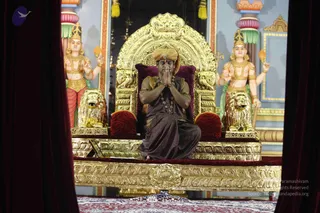 A seated figure in an ornate gilded throne with lion-armrest supports sits centrally between two gilded figures, with hands pressed together in namaste. He wears an orange-brown turban, a brown-maroon robe draped over one shoulder, and a beaded rudraksha-style mala with a pendant. The throne has red velvet cushions and a gold lion footstool, set before painted columns and a wall of blue, pink and gold, with maroon curtains framing the scene. Two golden standing figures with crowns hold lotus-like objects on each side. a person, appearance-based cues: orange turban and ornate mala in a throne setting. He is seated, hands in namaste.