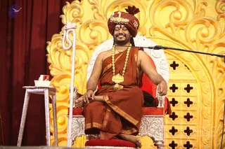 A smiling man in a maroon-red turban with a gold ornament and rudraksha-style adornments sits cross-legged on a white ornate throne with red cushions, in front of a gold carved backdrop with cross-shaped cutouts. He wears a saffron-brown dhoti and shawl and a heavy gold ceremonial necklace; a silver staff with a serpent-head top leans beside him and a small silver side table holds a cup. A microphone stand is at right.