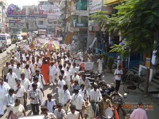 A large crowd walks down a busy Indian town street in a procession, mostly men dressed in white shirts and dhotis with ID lanyards, along with some women in pink and other saris. Near the centre, a woman in an orange-saffron sari walks at the front. Above the crowd, a large multicoloured umbrella with a yellow canopy is held aloft. Many people carry placards and banners, and a drummer in a khaki shirt beats a drum at the lower right. Parked motorbikes line the roadside, a white ambulance and cars travel the road, and shopfronts and signboards line both sides under a bright sky.