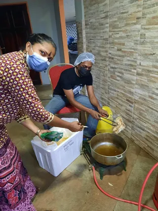 Photo of two volunteers preparing food in an indoor kitchen area with tiled wall. A woman in a red and cream patterned top and pink skirt, wearing a blue face mask and a bindi, holds a green packet over a white styrofoam cooler labelled in marker. A man in a black t-shirt and jeans, wearing a hairnet and black face mask, sits on a red upholstered chair and uses metal tongs over a large steel pot of oil on a portable gas burner with a red hose. A yellow jerrycan and foil sit nearby.