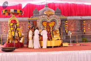Outdoor temple-style stage under a white and red canopy shows a seated figure on a gold throne decorated with a large heart-shaped backdrop of colourful beads and flower garlands. Several people in white dhotis and white shirts or saris attend the seated figure, with one man in an orange-red garment and a figure in a white dhoti and gold sash standing nearby, apparently performing a ritual offering. Flower garlands hang in yellow, white and red. A large black cooking vessel sits in the foreground, a golden lamp stand and a brass kalasha stand to the side, and a fan is visible on the right. Attendees appear to be performing abhishekam or puja. The seated figure's identity is not visually established.