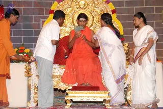 A seated figure in an orange-saffron robe sits on an ornate gold throne with a red cushion and red velvet backing, decorated with a garland of yellow, orange and red flowers. He holds a green leafy sprig or bundle near his face while a man in a white shirt and grey trousers bends toward him, and a woman in a white sari with lavender and gold border stands at his right, while a young woman in a white sari with gold-bordered pallu stands at the far right. At left, a man in an orange dhoti-style garment holds a tray of flowers and offerings. Gold lamps and white flower garlands decorate the platform on a brick-patterned wall.
