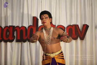 A performer in a stage dance costume stands before a white draped curtain with large red lettering behind him, partly reading 'ndat' and 'av'. He is bare-chested with multiple strands of white pearl necklaces, a green beaded collar, ornate gold armbands and bangles, and a gold-buckled waist belt over orange and purple dhoti-style lower garments. His face has red lip colour, dark eye makeup and a vertical tilak on the forehead. He holds his hands forward with fingers curled in a classical dance gesture. One person is visible.