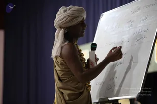 A man in a cream-white turban and a gold-tan draped garment, with a gold bead necklace, stands before a whiteboard and writes with a marker while holding a handheld microphone in his other hand. The whiteboard carries handwritten words including Energy, maya, sound, silence, undefined, dull and tamas in a grid. A blue curtain forms the backdrop.