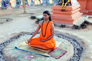A photo of a man seated cross-legged in meditation on a raised circular stone platform outdoors, bordered by white pebbles and decorated with white chalk kolam dots and a green painted vine pattern. He wears a saffron-orange unstitched robe, a long dark rudraksha mala with multiple strands, and a red bindi on his forehead, with long swept-back hair. His hands rest in a mudra on his lap, and a brass kalasha-style vessel sits beside him on a woven straw mat with a small maroon cloth. Behind him stands an orange-red peetha-like shrine base draped with marigold and jasmine garlands, with a deity murti partly visible behind a white cloth. Potted plants and a small pigeon are visible on the ground, and a line of stone statues and red signboards appears in the background. The man appears to be a probable Swamiji by appearance cues: long swept-back hair, saffron clothing and rudraksha mala, seated in meditation.