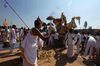 Outdoor ritual procession on dry dusty ground. In the foreground a man in white dhoti with an orange-gold border holds a white ceremonial vessel aloft. Behind him a decorated chariot with a canopy and garlands moves past, with orange-red umbrellas and a smoking fire pot. Many men in white shirts and white dhotis stand and move around the chariot and a long rope, and a gold trophy-like kalasha stands at left with red-white ribbons. Clear blue sky above.