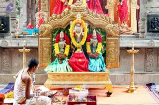 A golden ornate shrine houses three deity murtis garlanded with yellow and red flower garlands. The central murti holds a spear-like weapon and wears an orange cloth, flanked by two female murtis in green and turquoise sarees. A priest in white dhoti sits cross-legged at left, reciting before a microphone, with a fire pit of burning sticks, a tray of bananas and brass oil lamps around the shrine. Red and orange fabric drapes the background walls.