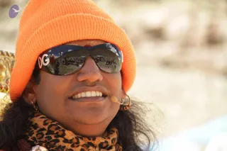 Close-up photo of a smiling man wearing a bright orange knit beanie, mirrored sunglasses with a "DG" logo on the temple, a small red tilak on the forehead, and a leopard-print shawl around his neck. A small clip-on headset microphone is visible near his cheek and gold hoop earrings hang from his ears. Reflected in the lenses are a hillside crowd and a wide open landscape under a blue sky. Background is soft-focus pale ground and a gold-patterned surface at left. Appearance-based cues such as the long hair and shawl are present, but the person cannot be confirmed, so the identity is unsure.