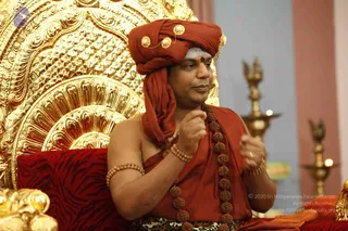 A man seated on an ornate gold throne with a large gilded backrest, wearing a deep orange-red turban adorned with gold and pearl-style ornaments and a red bindi on his forehead, with white ash marks above the brow. He wears an orange-red shawl over a bare shoulder, a long rudraksha mala with a matching beaded bracelet and armlet, and speaks while gesturing with both hands. He is seated on a red cushion with a tiger-stripe pattern. Brass oil lamps stand in the background against a pink and grey wall. Swamiji (probable) appears as the seated speaker, shown in orange robes, rudraksha beads and a turban.
