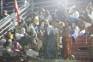 Night ritual photograph of a large black stone Shiva lingam decorated with a yellow marigold garland, being bathed with water. A man standing at the right, bare-chested with a rudraksha mala and wearing an orange dhoti, pours water from a brass kalasha over the lingam. Around the steps, many people sit in rows in white, yellow, purple and pink clothing, several holding small brass ritual vessels and offering bowls. Brass oil lamps stand on the steps at the right, and a red banner is visible on the left behind a metal railing.