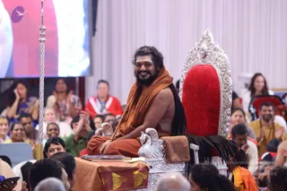 A man with long swept-back dark hair and a full beard, wearing an orange-saffron shawl over a bare chest with long dark hair flowing down his back, sits smiling on an ornate silver throne with a red velvet seat and carved lion-head armrests. A microphone is clipped to his face. A silver staff stands at left and a small table with a book and cloth lies in front. Seated audience members in saffron, blue and red clothing fill the hall, several clapping, and a large screen behind shows red imagery. a person appears in person, seated on the throne during a discourse-style gathering.