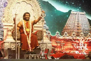 A seated man on an ornate silver throne with leopard-print cushions, wearing a rust-orange robe with gold trim, a long flower garland, a gold pendant necklace and a rudraksha-style bracelet, smiling and raising a silver trident-like staff in his right hand. Behind him a large backdrop shows a starry night sky over a red-tinted gopuram temple tower and a deity murti. Small brass oil lamps and offerings sit on the stage below. The seated figure is the only visible person.