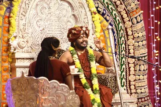 A seated man in a saffron-orange turban and saffron robe, wearing a rudraksha mala and a multi-strand gold necklace and a flower garland of yellow and green blooms, smiles and raises one hand while speaking into a microphone on a gold-and-jewelled throne. Facing him, in the left foreground, a woman with long dark hair pulled back, wearing a rust-red top, sits in a silver chair. Behind them, the ornate throne backdrop is covered in silver filigree, gem-studded panels and gold embroidery, with strings of fairy lights at right. Appearance-based label: a person, seated on the throne in the centre; the caption is not visible here, so no name is printed in the frame.