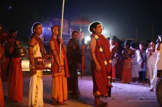 Night outdoor procession at a dusty ground lit by a bright floodlight and blue-white light. A woman in a rust-red saffron robe and a long multicoloured flower garland, red, white and yellow blooms with a red tassel, stands with her eyes lifted and hands held near her body, ready at the front. To her left, a man with a shaved head and a red-and-white shawl holds a tall golden staff, and a man in white dhoti with gold border and a red sash carries a brass pot topped with flowers. Behind them, a person in orange robes and a red-and-white shawl stands near a pole, and a crowd of devotees in white and red garments stands at the edge, some with hands folded. A large umbrella and a decorated float or structure are visible in the background.