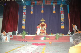 A stage decorated for a Valentine's Day celebration with balloons and rose petals. A man seated on an ornate gold-and-red throne with a golden canopy above is surrounded by balloons; he wears an orange robe. A woman in white with an orange shawl stands beside him holding a red heart-shaped balloon. Colourful painted cloth hangings with elephant motifs hang from the purple backdrop. Seated women in white and orange, and a few men in orange, sit on the sides of the stage and the floor, with palm fronds and a fan visible.