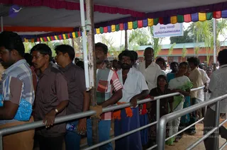 A photograph of devotees queuing under a multicoloured tent canopy at a Karthikai Deepam event, with a long line of people waiting behind steel barrier railings. A man in the foreground wears a striped shirt and carries a blue plastic bag, while a bearded man in a white shirt and orange-and-blue lungi stands at the rail. A young girl in a dark dress and a woman in a green sari stand to the right. A Tamil sign hangs above the queue, and palm trees and a red-roofed building appear behind. No a person candidate is clearly visible in this frame.