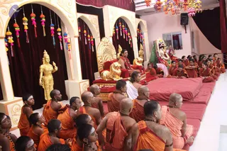 Devotees in saffron and orange robes sit on red floor cushions in a decorated hall during a satsang. At the centre a figure in red and gold robes with a garland sits on a large golden throne flanked by golden lion figures. Behind and to the right, a deity murti is dressed in green and red with a crown and garlands. Several shaved-headed devotees in orange wear white tilaka and rudraksha malas in the foreground. A golden standing deity figure stands at the left beneath arches hung with colourful tassels, and paper lanterns hang from the ceiling.