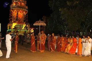 Night procession on a dirt ground lit by strings of green fairy lights in a tree. A tall illuminated ratha/chariot with a red and gold shrine and carved figures stands at left. A man in a red-orange robe with a red turban and a long beaded necklace holds a large ornate parasol over a line of devotees; a second man in a saffron robe holds a staff-like rod beside him. Women in orange and red saris and men in white dhotis walk in a line, several with hands folded in greeting. A cameraman in white stands at left filming. Shown as a temple procession with a chariot and parasol.