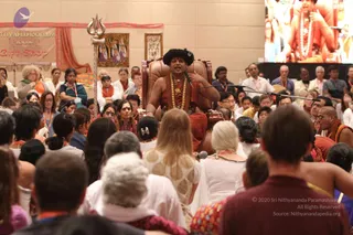 A seated man in a dark fur-trimmed cap, red-orange robe and gold-beaded necklaces sits on a brown leather throne behind a microphone stand, gesturing with his hand while speaking to a large seated audience. A silver trident stands to his left in front of a banner reading Nithyanandam 1008 Gift Shop. A large screen above shows a close-up of a figure in orange with rudraksha beads. Audience members in white, orange and saffron clothing fill the foreground and rows behind, many wearing orange lanyards. a person is seated on the throne, identified by the fur-trimmed cap, gold necklaces and orange robe, and is the central speaker.