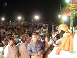 Night outdoor gathering with a very large crowd of people standing and seated before a raised platform lit by overhead lamps. In the right foreground, a figure in a saffron-orange turban and orange robe with a long rudraksha-style bead necklace stands at the platform edge, facing the crowd; the orange canopy above is decorated with green banana stalks and hanging fruit. Men in white kurtas and dhotis and people in sarees and salwar kameez fill the middle ground, while a man in a blue checked shirt is blurred in the foreground. Lamps and a dark tree line frame the scene.