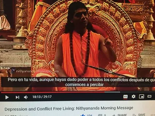 A screenshot of a YouTube video player showing a man in orange sleeveless robes with a long rudraksha mala speaking into a microphone in front of an ornate gold throne with carved floral arches. Behind him are brass oil lamps and a red cloth. A Spanish subtitle overlays the lower frame, and the video title below reads Depression and Conflict Free Living: Nithyananda Morning Message, with a progress bar at 18:13 of 29:17.