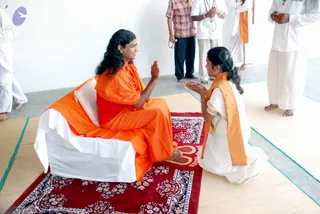 Indoor photograph of a long-haired man in an orange saffron robe seated on a white-covered cushioned seat atop a red patterned carpet, raising one palm toward a woman kneeling before him. She wears a white sari-style dress with an orange shawl over her shoulders and her hands are pressed together in a gesture of respect. Several people stand around the room in white clothes, some in orange shawls, one in a plaid shirt, some holding rudraksha malas. The man appears to be a person, identified by his long hair and saffron robe on a throne-like seat, with the woman devotee kneeling in front of him in darshan. A woody mat covers the floor and a faint purple logo appears in the upper left corner.