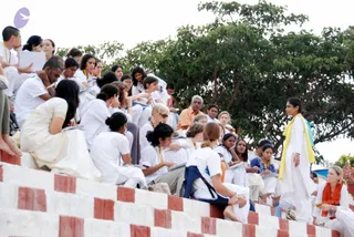 Outdoor photograph of a seated group of participants on red-and-white tiered steps beneath a large leafy tree. Most wear white clothing, many with yellow or orange cords and some holding notebooks and pens, appearing to listen or take notes. A woman in a white outfit with a yellow shawl stands at right, facing the group, while a man in an orange shirt sits in the middle of the crowd. Other participants sit in the foreground, one in a white dress and another in a blue-and-white patterned top. A blonde woman with an orange scarf sits at far right.