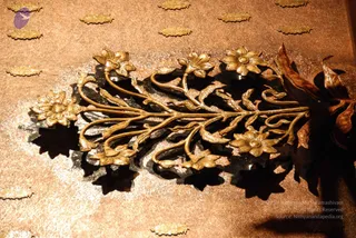 Close photograph of an ornate golden floral decoration. The gilded piece has a central stem curling into multiple blossoms, with lotus-like flowers and leaves rendered in intricate relief. Warm lighting casts dark shadows beneath the ornament. A faint circular emblem appears at the upper left corner. No people are visible.