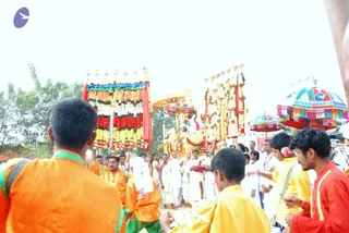 Outdoor procession in daylight with a decorated palanquin-style float carried through a dusty open area. Two tall flower-garland panels with red, yellow and white blossoms flank a central golden canopy under which a figure in a white dhoti sits. Devotees in saffron and orange robes walk alongside, with several men in yellow and red shirts in the foreground. Men in white dhotis and white shirts follow the float, some with white shawls. Multicoloured umbrellas with tassels stand at the right and a green tree appears at the left.
