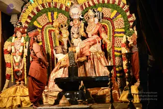 Photograph of a temple sanctum scene at night, lit by warm lamps. Centre-right, a deity murti pair seated on a throne-like platform: a crowned figure with a trident-like staff and a seated female figure in red and gold silk, both adorned with heavy gold jewellery and white crowns. A black shivalinga stands on a black stone pedestal before them. The arch behind is densely decorated with garlands of marigold, rose and multicoloured flowers in red, yellow, purple and white. On the left, a standing male figure in saffron-orange robes with a red headband and long dark hair, hands folded at his chest, stands beside a flower-garlanded figure in gold. Brass oil lamps and a small brass figure sit in the foreground. Two statues are the only other figures; no living people are clearly visible beyond the saffron-robed man.