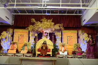 A stage with a golden ornate throne centred between two gold-trimmed chairs, with a gilded tree-like decoration above. Five people are seated on the stage: a man in orange robes on the far left, a man with a green shawl beside him, a central figure in saffron-orange robes with a rudraksha mala seated on the golden throne, a man in white with an orange shawl, and a woman in a purple sari. A woman in a magenta sari holding a microphone stands at the right edge. Red curtains hang behind, with a trident and golden statuary on the sides.