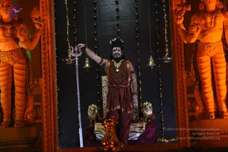 A figure stands in an illuminated shrine-like alcove between two large orange-lit carved guardian statues with raised arms. The central figure wears a maroon and red-brown wrap and dhoti, a gold necklace over layered rudraksha malas, a gold crown and a painted face. He grips a silver trident with one hand. Beside him are gold lion heads on a dark cushioned throne draped with striped cloth, a brass oil lamp, hanging bells and strings of beads and lights against a black backdrop. a person appears in the central position, shown in a ceremonial seated setting with clear ornate cues. No other people are visible.