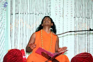 A seated man with long dark hair and a red tilak on his forehead sits cross-legged on a red zebra-striped cushion in front of a white curtain hung with dark and orange beaded strands. He wears an orange sleeveless robe and a long gold-beaded necklace, with his palms open and his eyes raised upward as if chanting or singing. A microphone on a flexible stand is beside him, and a pink book rests on a wooden book stand in front of him. Two red tambourine-like percussion instruments sit at the sides. a person: long swept-back hair, saffron robe and ornate gold necklace, seated in the role of a speaker or singer leading a satsang-style performance.