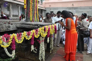 A photograph at a temple courtyard showing a figure with long dark hair in a saffron-orange sleeveless robe and matching orange garment, barefoot, standing with palms pressed together in namaskar before a dark stone structure draped with long garlands of yellow, pink, and white flowers. Behind them, a crowd of devotees in white shirts and dhotis watches, and a photographer with a camera stands at right. Pink temple walls and white pillars surround the scene. The figure matches the appearance cues of the a person, so the identity is noted here as a person, shown in a worship posture; the mood is reverent.