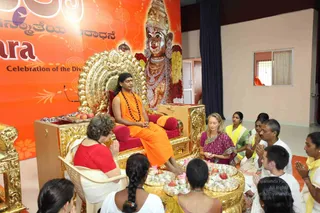 A seated figure in saffron-orange robes and a rudraksha mala sits on an ornate gold throne with red cushions, feet resting on a gold footstool, as people kneel and sit on the floor before him. Behind the throne stands a large golden deity murti adorned with pearl necklaces and flower garlands. Devotees in white and yellow clothing press their palms together in namaskar. A woman in a magenta sari sits cross-legged on the right, and an older woman in red sits in a chair on the left. Silver-foiled trays of sweets and fruit with flowers sit on a gold stand in front. An orange banner with Kannada text and a decorative logo hangs behind the throne, and the setting is a bright indoor hall with cream walls and dark curtains. The figure's appearance is consistent with the page's reference to a person, presented in a seated satsang setting.