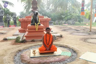 A seated man in saffron-orange robes and a rudraksha mala sits cross-legged in meditation on a small saffron-cushioned seat over a rangoli-decorated circle lined with grey gravel. He wears a red tilak on his forehead, hands folded in his lap. Behind him stands a large black Nandi bull statue garlanded with yellow marigolds atop a red stepped pedestal, with grey stone deity statues and palm trees in the background. A brass kalasha sits beside him on a woven mat, and tall decorative banners in orange, white and blue frame the right side of the scene. Sunlight filters through the palms over an open dirt courtyard.