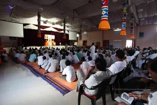 A large hall filled with seated devotees in white and light clothing, many sitting on orange and blue woven mats on the floor and others on black chairs, facing a raised stage at the far end. On the stage, a figure in orange sits on a golden throne-like seat beside another person in white, with a large screen above showing a close-up image of a seated figure in orange. Tall decorative cylinders with orange tassels and colourful patterns hang from a white draped ceiling. A standing man in white stands near the stage, and a projector screen flanks the stage. Some attendees hold booklets.