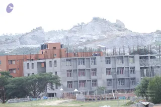 A photograph of an unfinished concrete multi-storey building under construction at a campus site, with a rocky, quarried hillside behind it. The grey concrete frame has rows of windows with red shutters, and rebar rods stick up from the upper floors. Orange-plastered wings and black water tanks stand on the left. Two workers are visible on the upper roof, and a fenced lawn with a few trees and green direction signs runs along the front. The rocky hill is pale and sparsely vegetated.