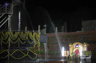 Night photograph of an outdoor abhishekam at a large black stone shivalinga wrapped in yellow and green marigold garlands, with water streaming down its sides. To the right, a seated figure in orange-red robes sits in an arched shrine decorated with yellow and green flower garlands, lit by two white lamps, with a small green-clad attendant beside the throne. Brick terraced wall with plants behind, and a metal staircase at left. The water pool holds floating garlands.
