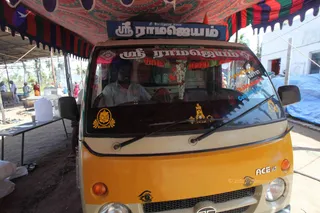 A yellow Tata Ace mini truck parked under a colourful striped canopy at a relief camp, its windscreen and roof sign reading ශ්‍රී රාමജെයം in Tamil script with garlands of orange and white flowers hanging across the windscreen. A man in a white shirt sits at the wheel. Stickers of a lion emblem and a small temple-like figure decorate the windscreen. Behind the vehicle, a steel vessel, a water container and a table are set up on dirt ground under awnings, with a few people in the background.