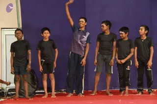 Six boys stand in a line on a red stage floor in front of a blue backdrop, performing a culturals act. They are barefoot and wear dark t-shirts and shorts or track pants, with a few in a purple-and-navy athletic top. Several have light markings on their faces and arms, and the boy in the centre raises one hand overhead. A keyboard and audio cables sit at the left edge of the stage. Scattered petals or confetti lie on the red floor.