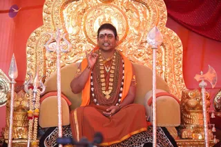 A seated man in orange and saffron robes sits on a cushioned golden throne with ornate gilded carvings, flanked by silver tridents and spears and a golden lion-form armrest. He wears a long multi-strand rudraksha mala, a heavy gold necklace, bangles and a white-and-red tilak on his forehead, and raises one hand in a teaching gesture while speaking into a head microphone. Red draped fabric forms the backdrop. a person: a man with a sweeping dark hairstyle and ceremonial gold necklaces and saffron robes sits on the throne delivering a satsang discourse.