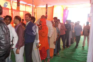 A photograph of a long queue of men inside a tent with orange and yellow fabric walls and a green mat floor. In the foreground a man in an orange hooded robe and red dhoti stands in line, holding a small yellow object. Others wear jackets, collared shirts, kurtas and sweaters, some barefoot. At the far right a framed portrait hangs on the tent wall beside the queue. No candidate for a person is clearly identifiable by appearance, so the person is described by role only.